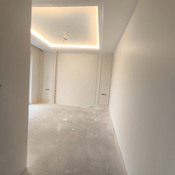 MFI Mateusz Lisczyk - Białe wnętrze pokoju z dekoracyjną sztukaterią ścienną i podświetlanym sufitem kasetonowym. Surowa podłoga przed wykończeniem. Widok z perspektywy wejścia.