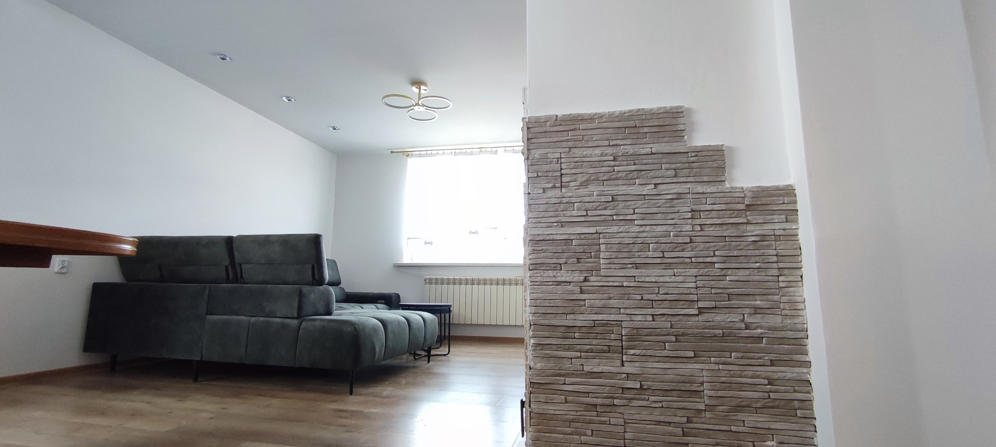 Salon z szarą sofą i ścianą wyłożoną dekoracyjną, beżową cegłą. Widoczne białe ściany i drewniana podłoga, nowoczesna lampa sufitowa.