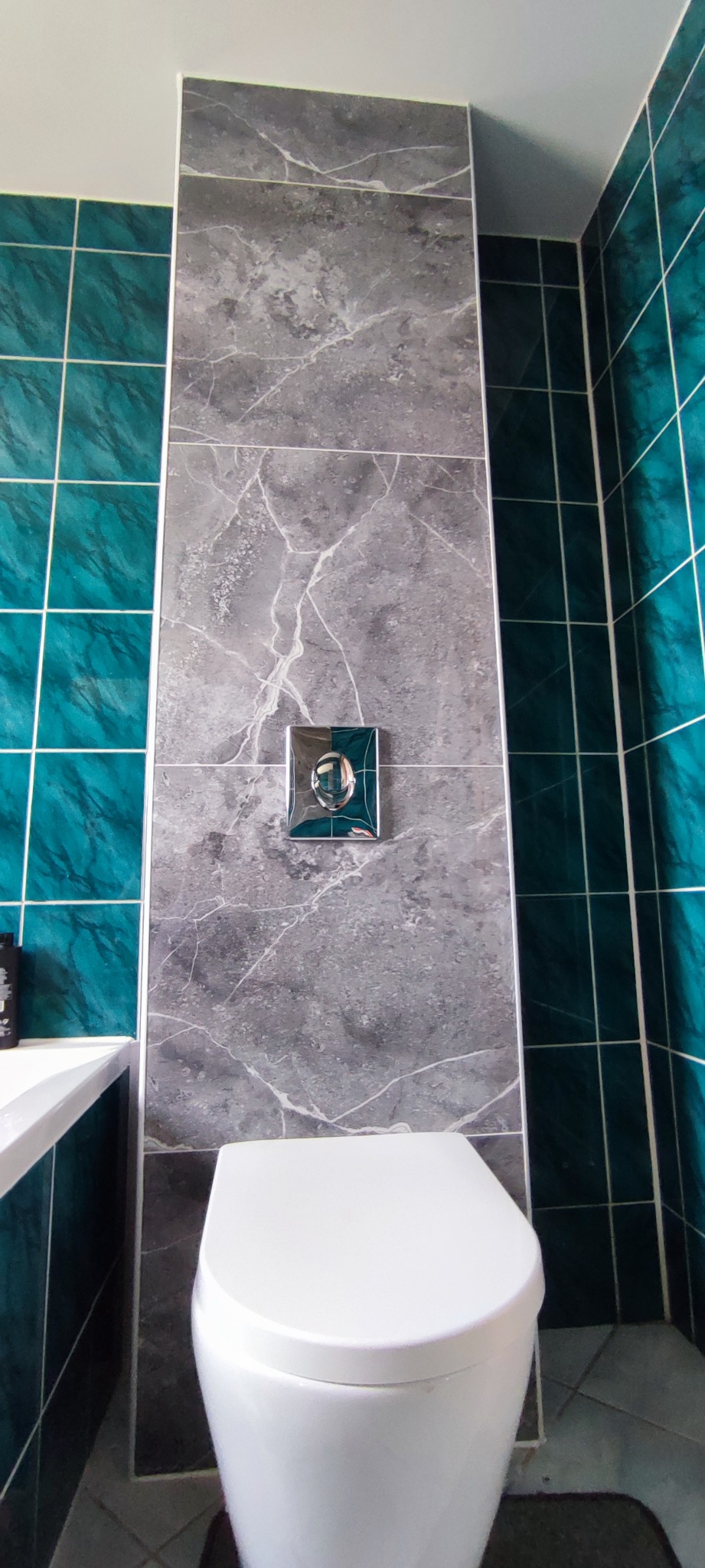Nowoczesna toaleta z szarą płytką imitującą marmur i turkusowymi akcentami. Widoczny fragment wanny. Czyste, wykończone wnętrze łazienki.