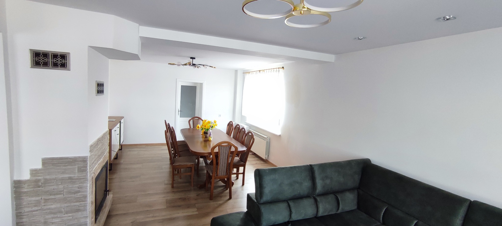 Jasne wnętrze z drewnianą podłogą, stołem i szarym narożnikiem. Ściany białe, oświetlenie sufitowe. Widoczny kominek z okładziną imitującą kamień.