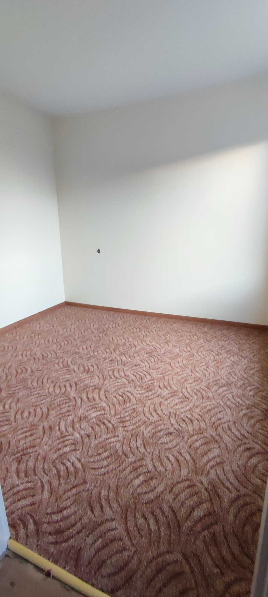 Pokój po remoncie: kremowe ściany i wzorzysta, brązowa wykładzina dywanowa. Widoczny fragment listwy przypodłogowej i gniazdko elektryczne na ścianie.