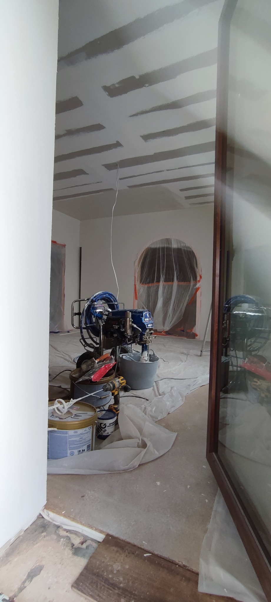 Wnętrze w trakcie prac wykończeniowych: sufit z płyt kartonowo-gipsowych, sprzęt do malowania natryskowego, zabezpieczone folią okno i drzwi. Widoczne narzędzia i farby.