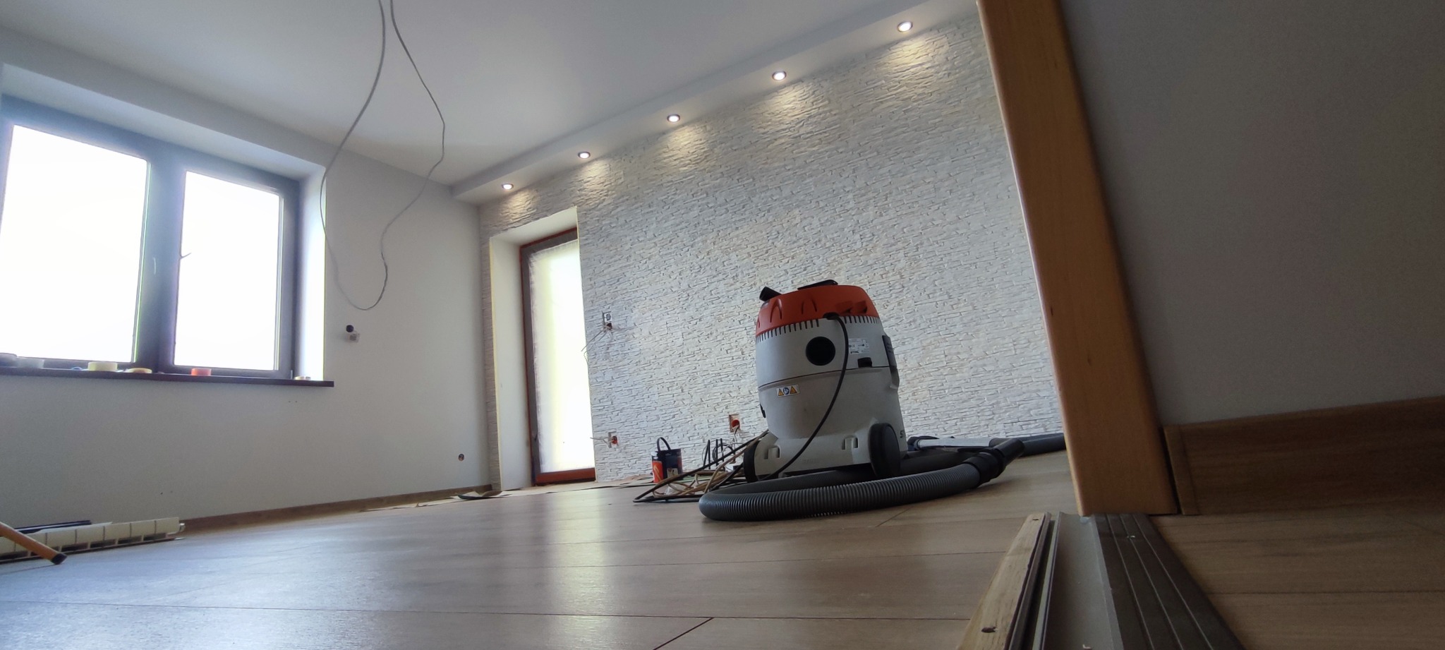 Pokój po remoncie z ułożonymi panelami podłogowymi, widoczny odkurzacz przemysłowy i ściana z dekoracyjną strukturą. Widoczne listwy przypodłogowe i oświetlenie sufitowe.