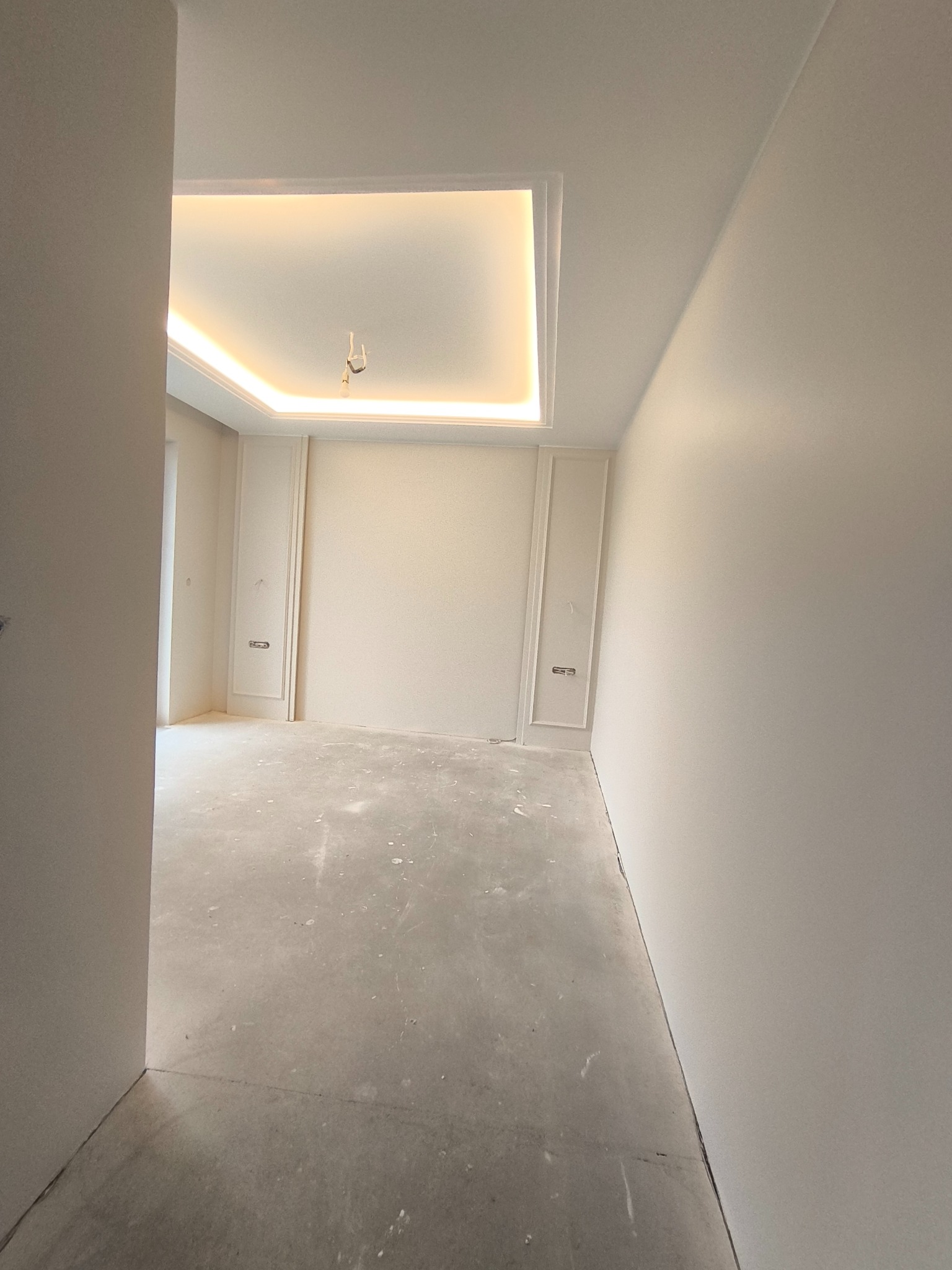 Białe wnętrze pokoju z dekoracyjną sztukaterią ścienną i podświetlanym sufitem kasetonowym. Surowa podłoga przed wykończeniem. Widok z perspektywy wejścia.