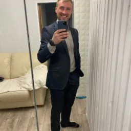 Uśmiechnięty mężczyzna w garniturze robi selfie w lustrze, widoczna sofa i drewniana boazeria.