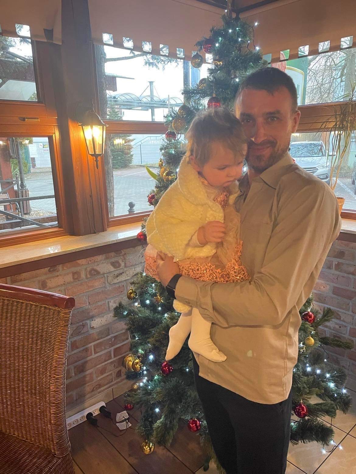 Mężczyzna trzyma na rękach małą dziewczynkę na tle choinki w pomieszczeniu z ceglaną ścianą i dużym oknem z widokiem na zewnątrz.
