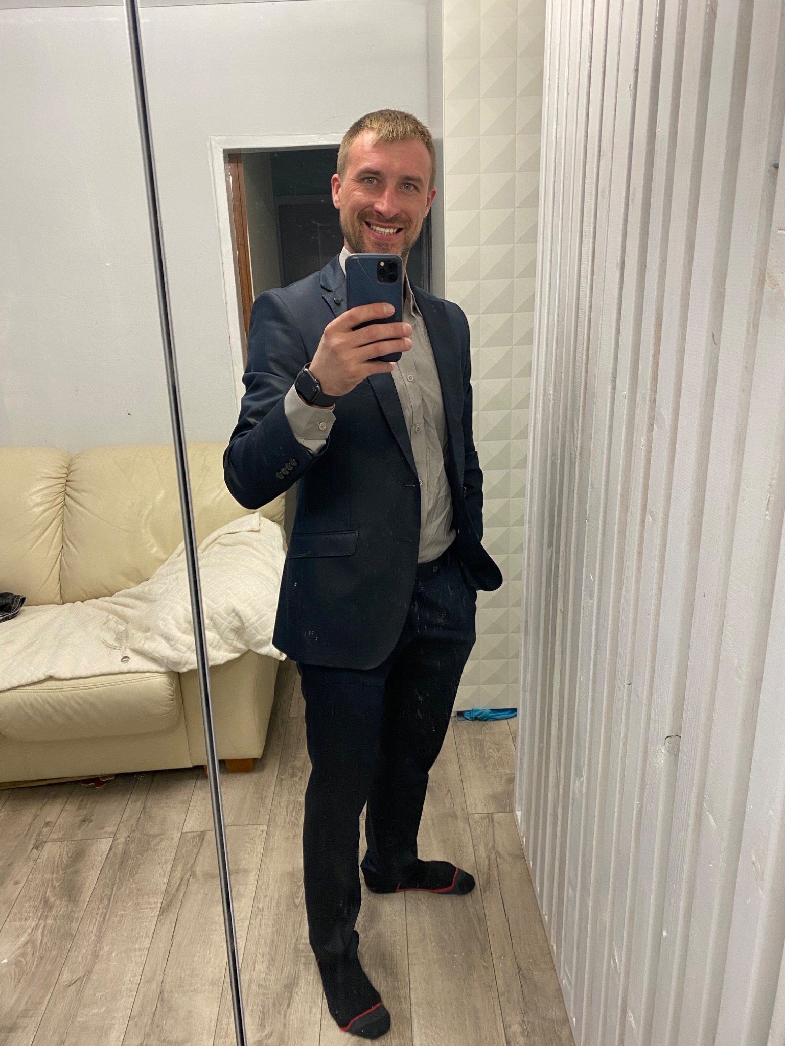 Uśmiechnięty mężczyzna w garniturze robi selfie w lustrze, widoczna sofa i drewniana boazeria.