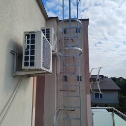F.H.U Gabriel Kowalski - Dwa klimatyzatory zamontowane na elewacji budynku obok metalowej drabiny ewakuacyjnej z zabezpieczeniem klatkowym, na tle błękitnego nieba z chmurami.