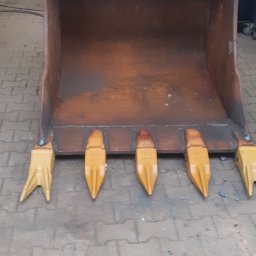 F.H.U Gabriel Kowalski - Pomarańczowo-brązowa łyżka koparki z żółtymi zębami, stojąca na brukowanej powierzchni w warsztacie.