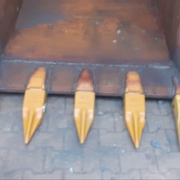 F.H.U Gabriel Kowalski - Metalowa łyżka koparki z przyspawanymi, żółtymi zębami, widoczne ślady spawania i rdzy, na tle szarej kostki brukowej.