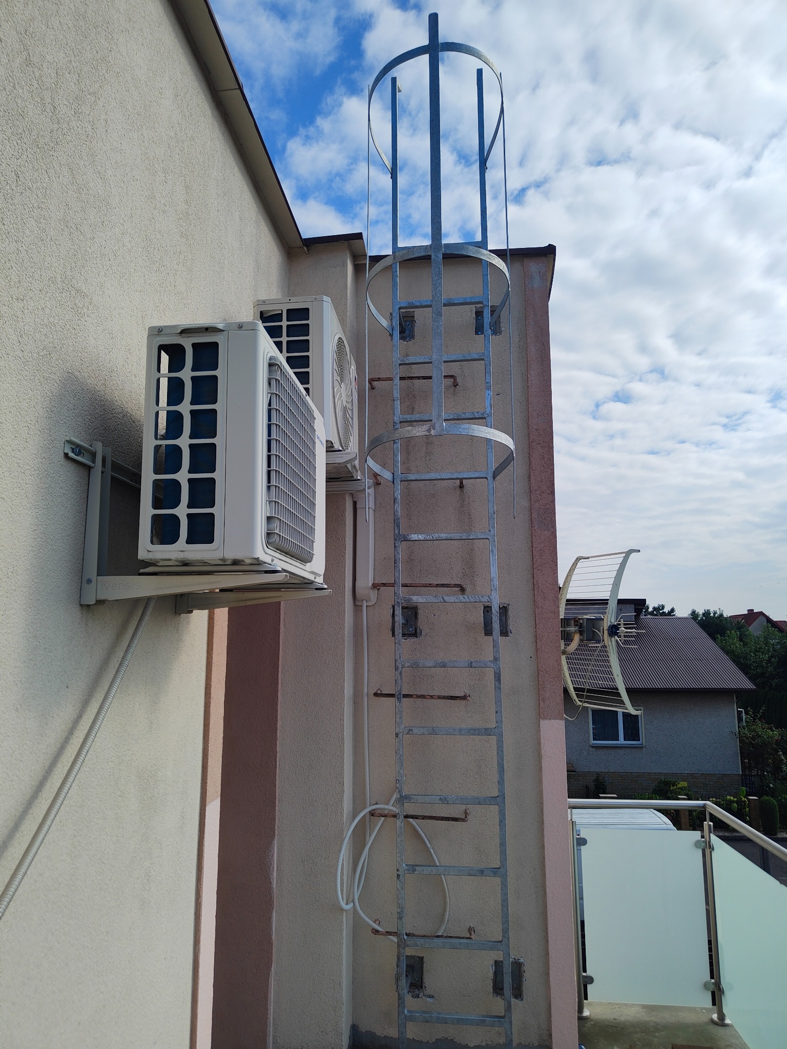Dwa klimatyzatory zamontowane na elewacji budynku obok metalowej drabiny ewakuacyjnej z zabezpieczeniem klatkowym, na tle błękitnego nieba z chmurami.