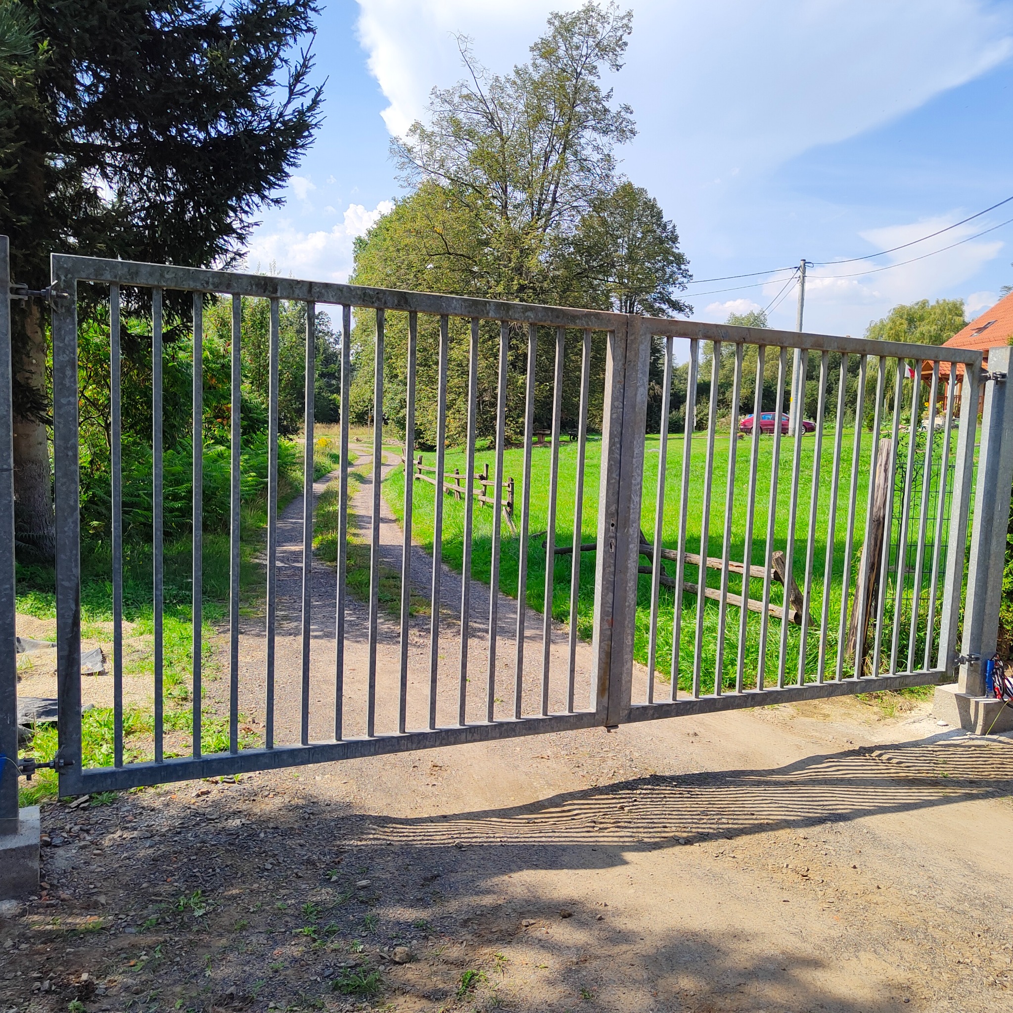 Otwarta, metalowa brama wjazdowa z pionowymi szczeblami, prowadząca na wiejską drogę z widokiem na zieleń i drzewa w oddali, jasne światło słoneczne.