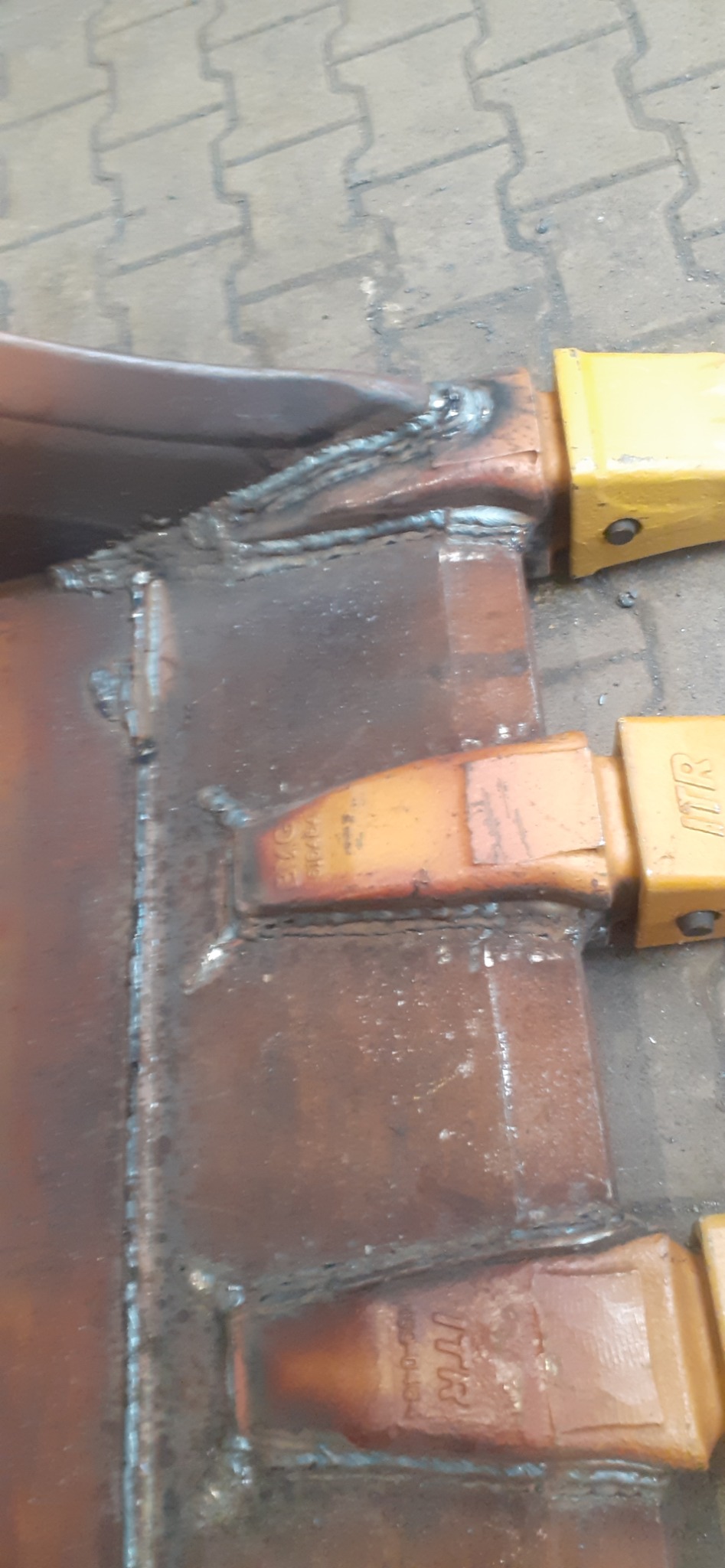 Spoina spawalnicza łącząca ząb łyżki koparki z korpusem, widoczne ślady użytkowania i oznaczenia producenta ITR na zębach.