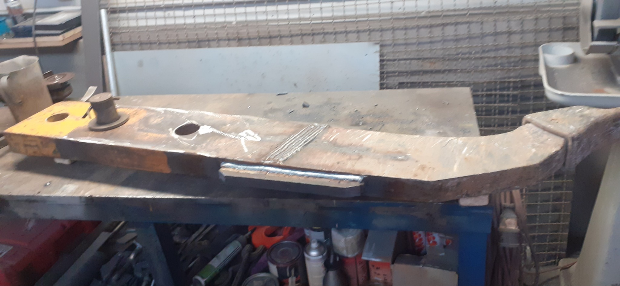 Żółto-brązowy element metalowy z widocznymi śladami spawania leżący na stole warsztatowym, w tle narzędzia i akcesoria warsztatowe.