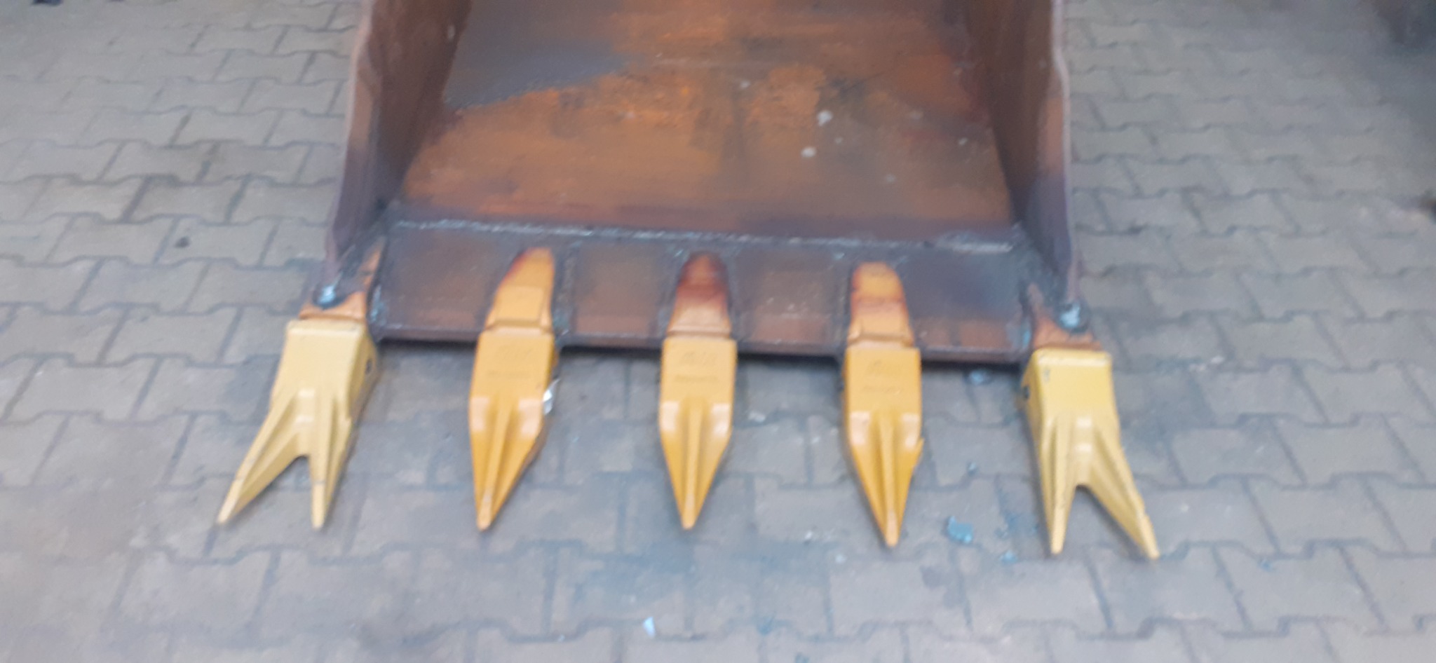 Żółte zęby łyżki koparki przyspawane do brązowej krawędzi łyżki, widoczne ślady spawania, tło z szarej kostki brukowej.