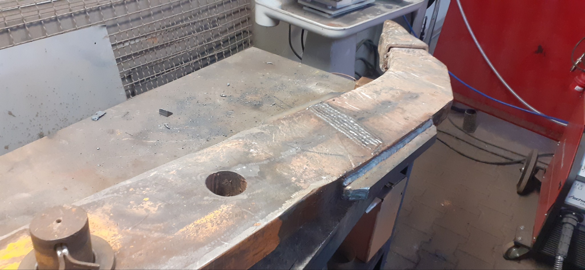 Ciężki, metalowy element konstrukcyjny z widocznymi śladami łączenia spoinami, leżący na warsztatowym stole; widoczne otwory montażowe i ślady obróbki.