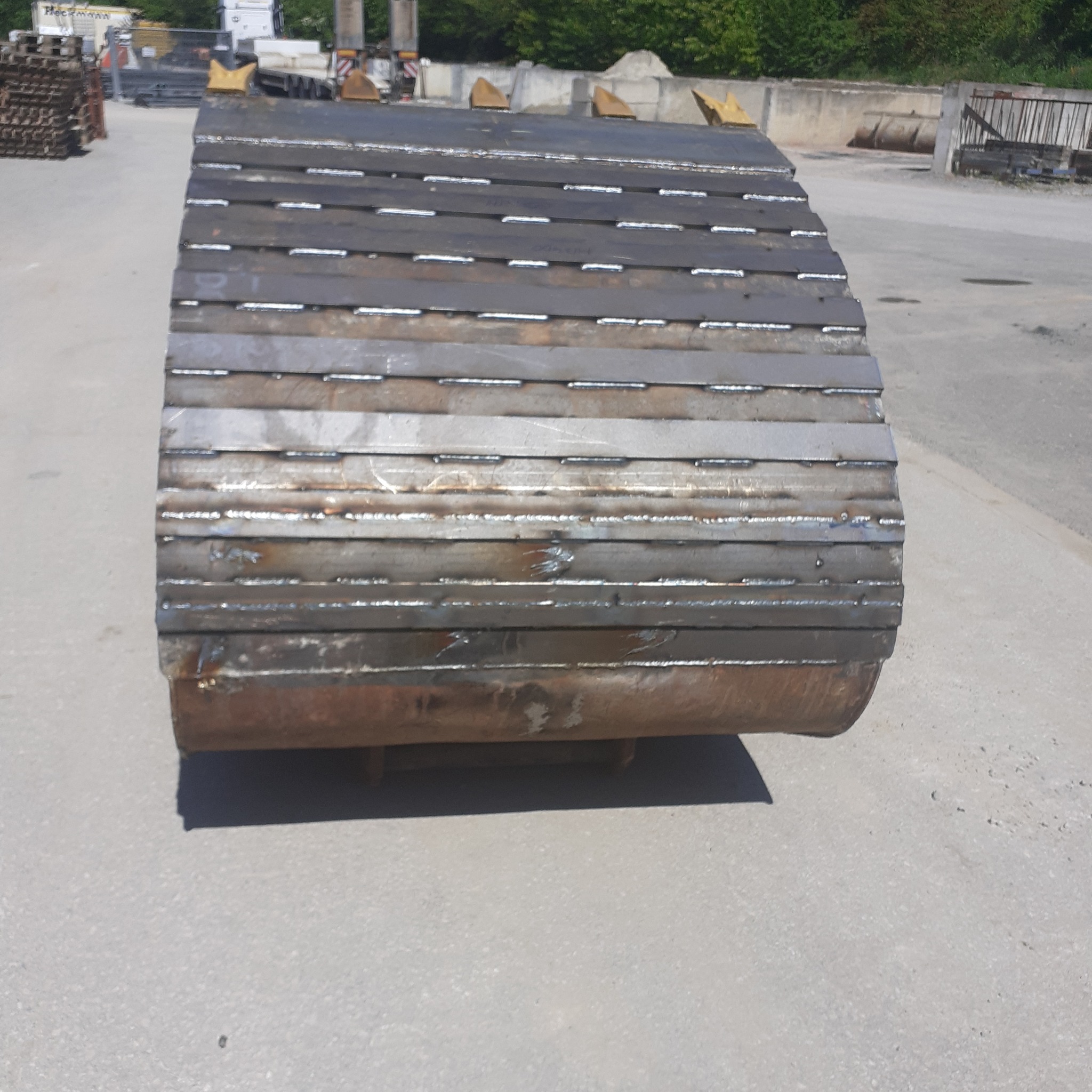 Metalowy walec z przyspawanymi poprzecznymi płytami, widoczne ślady spawania, maszyny budowlane i składowane elementy w tle.