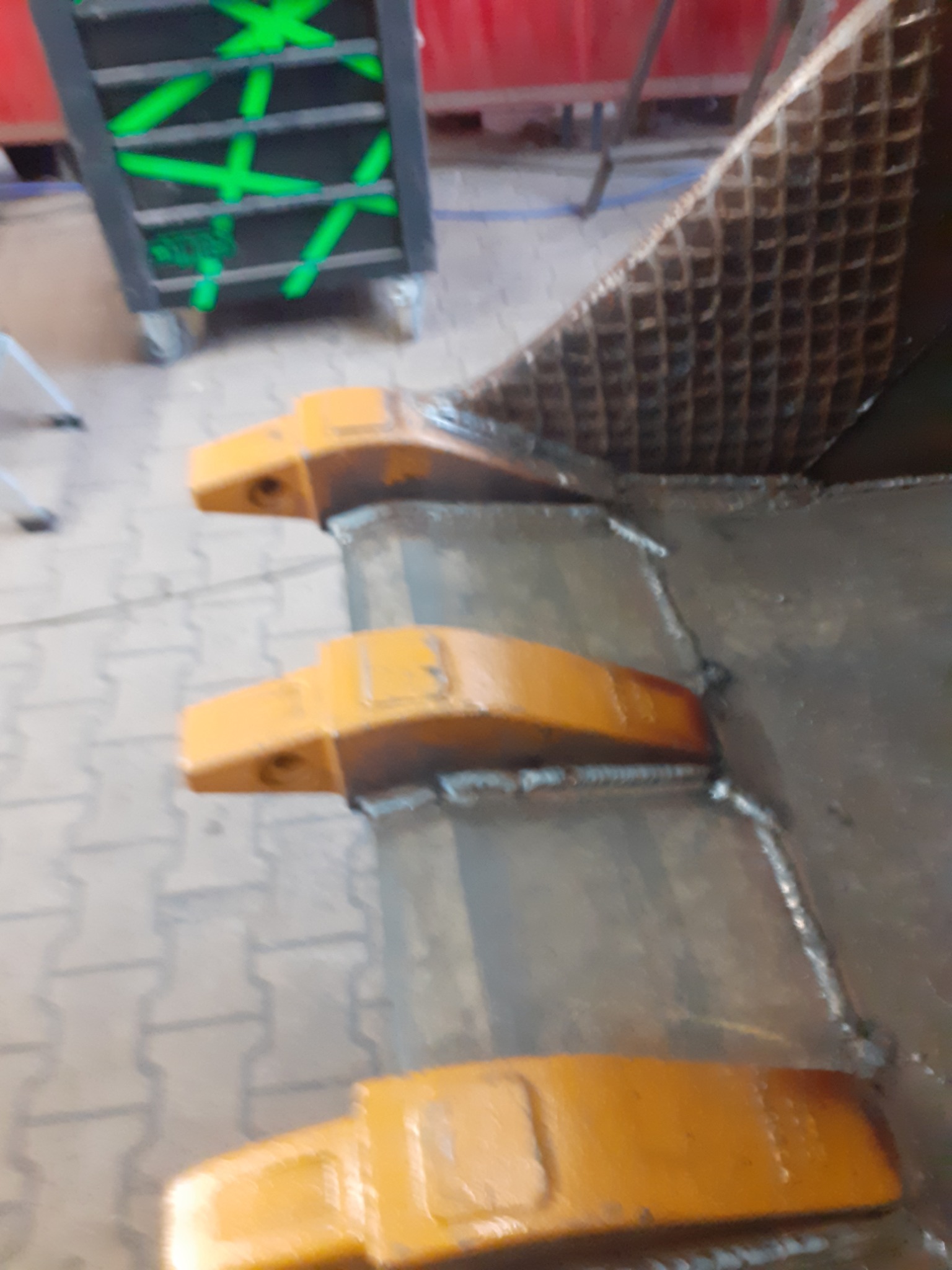 Żółte zęby łyżki koparki przyspawane do stalowej konstrukcji, widoczne spoiny i narzędziowy wózek w tle.