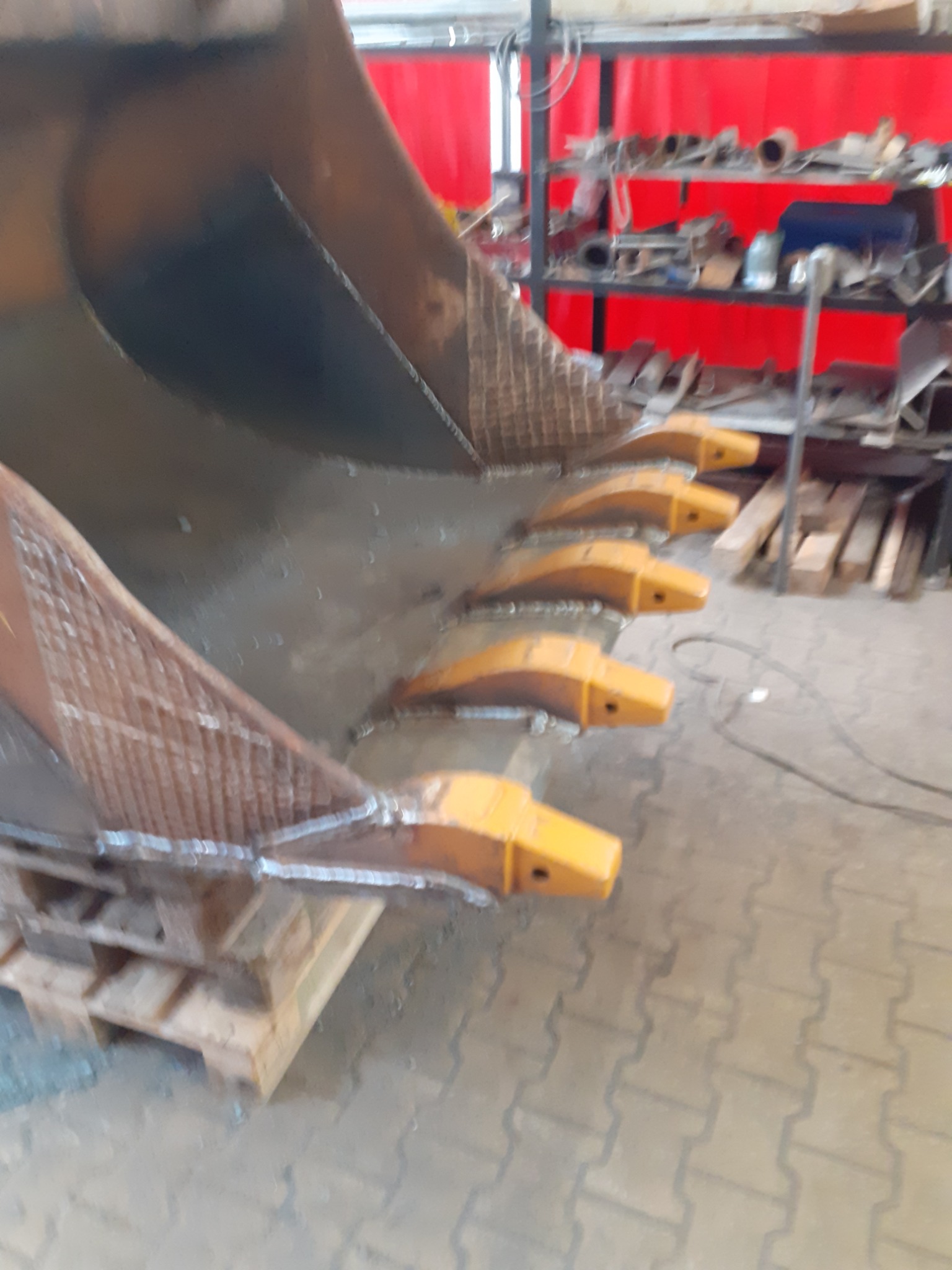 Ciężka łyżka koparki po wymianie zębów, widoczne nowe, żółte zęby i ślady świeżych spawów, całość umieszczona na palecie w warsztacie.