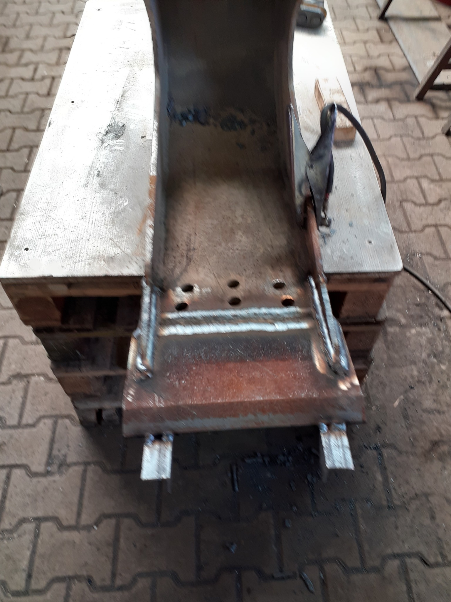 Metalowa konstrukcja po spawaniu, umieszczona na drewnianej palecie, widoczne spawy i ślady korozji, w tle kostka brukowa.