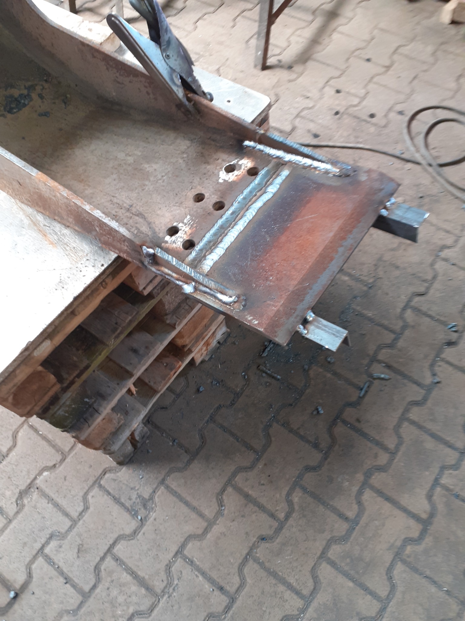 Metalowa konstrukcja z widocznymi spawami, umieszczona na drewnianej palecie, z zapiętym zaciskiem; tło stanowi chodnik z kostki brukowej.