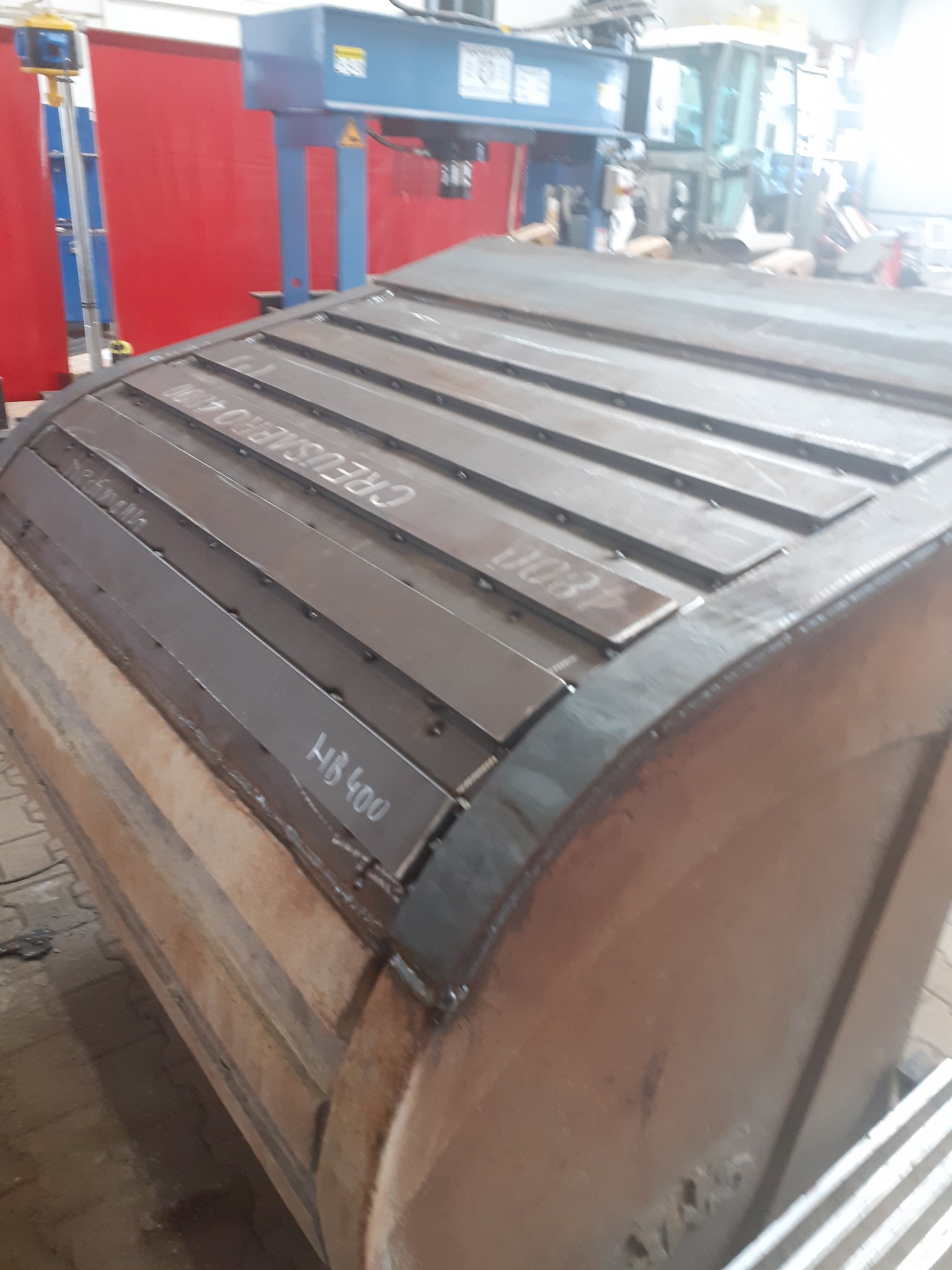 Częściowo zespawany, zakrzywiony element konstrukcyjny z blachy, z przyspawanymi wzmacniającymi paskami. Widoczne napisy 'CREUSMEIRO 4100' oraz 'HB 400'. W tle widoczna spawarka automatyczna.