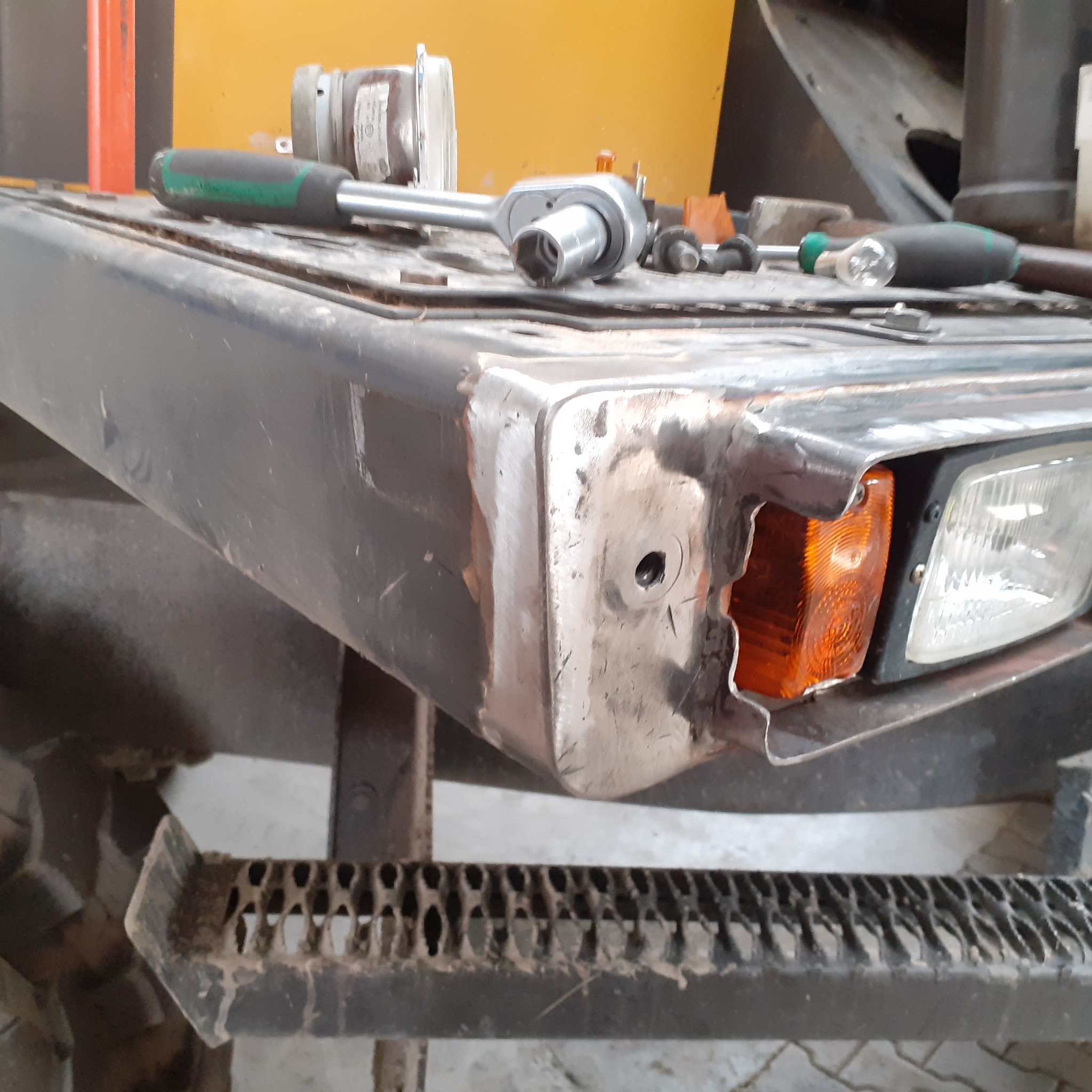 Naprawiony narożnik metalowej ramy pojazdu z widocznymi śladami spawania i szlifowania, narzędzia leżące na górze, w tle żółta maszyna.