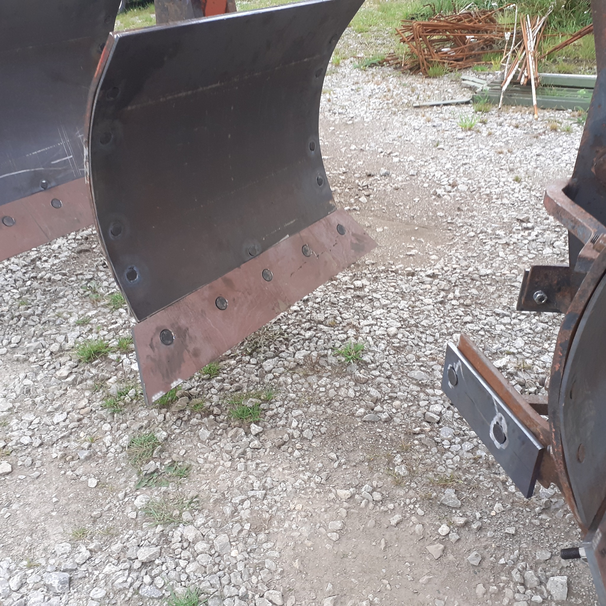 Metalowe lemiesze do pługa z widocznymi śladami spawania i mocowania śrubami, leżące na żwirowym podłożu.