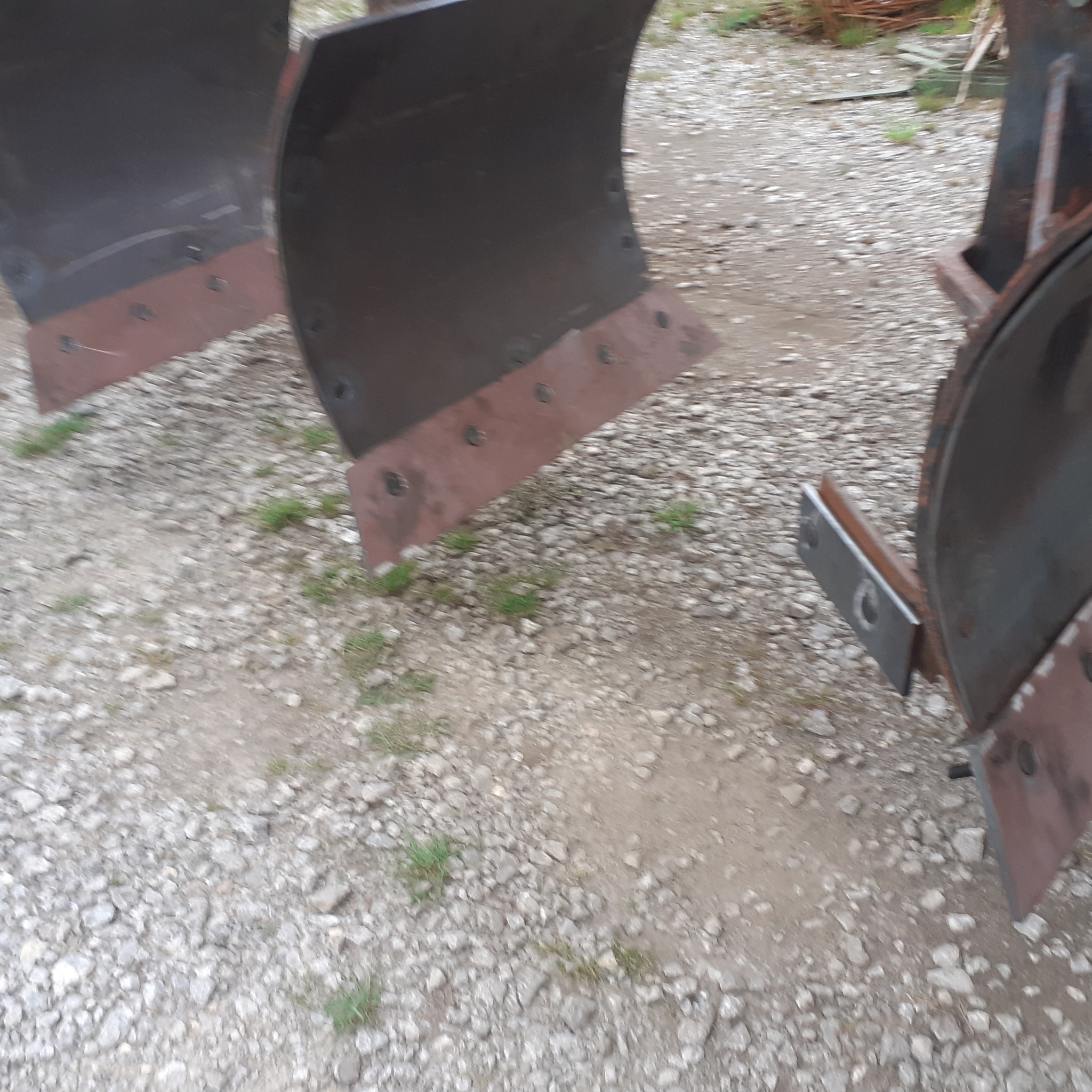Części metalowe do maszyny rolniczej, prawdopodobnie lemiesze pługa, z widocznymi śladami spawania i mocowania na tle żwirowego podłoża.