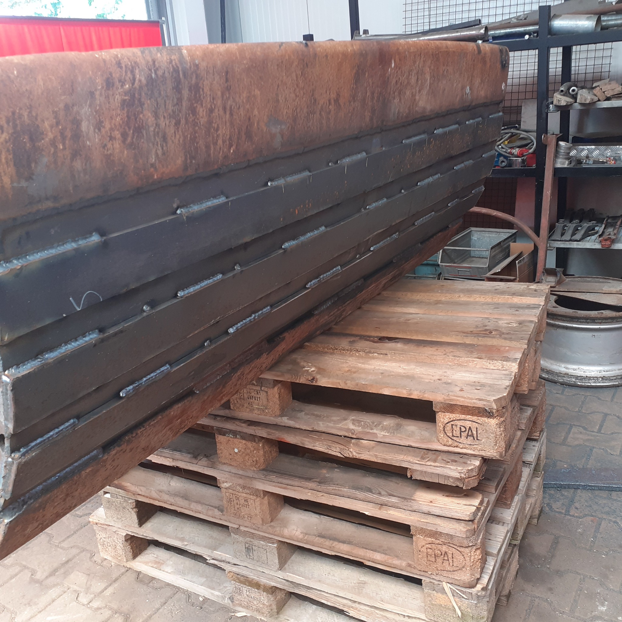 Ciężka, zespawana konstrukcja z metalowych płyt z widocznymi spawami, ułożona na stosie drewnianych palet z oznaczeniem EPAL w warsztacie.