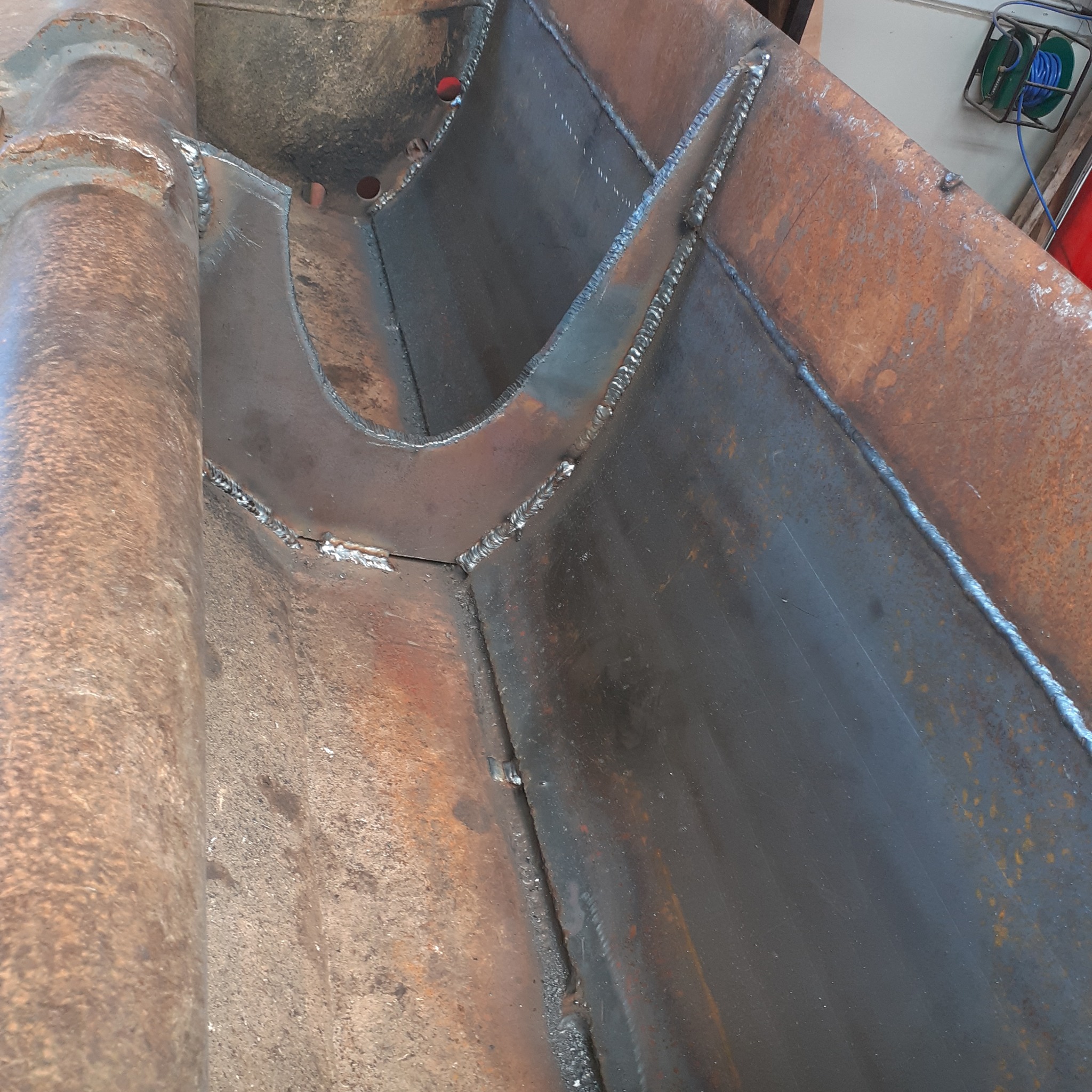 Wewnętrzny widok konstrukcji metalowej z widocznymi spawami łączącymi elementy, rdzawy nalot na powierzchni metalu, przewody elektryczne w tle.