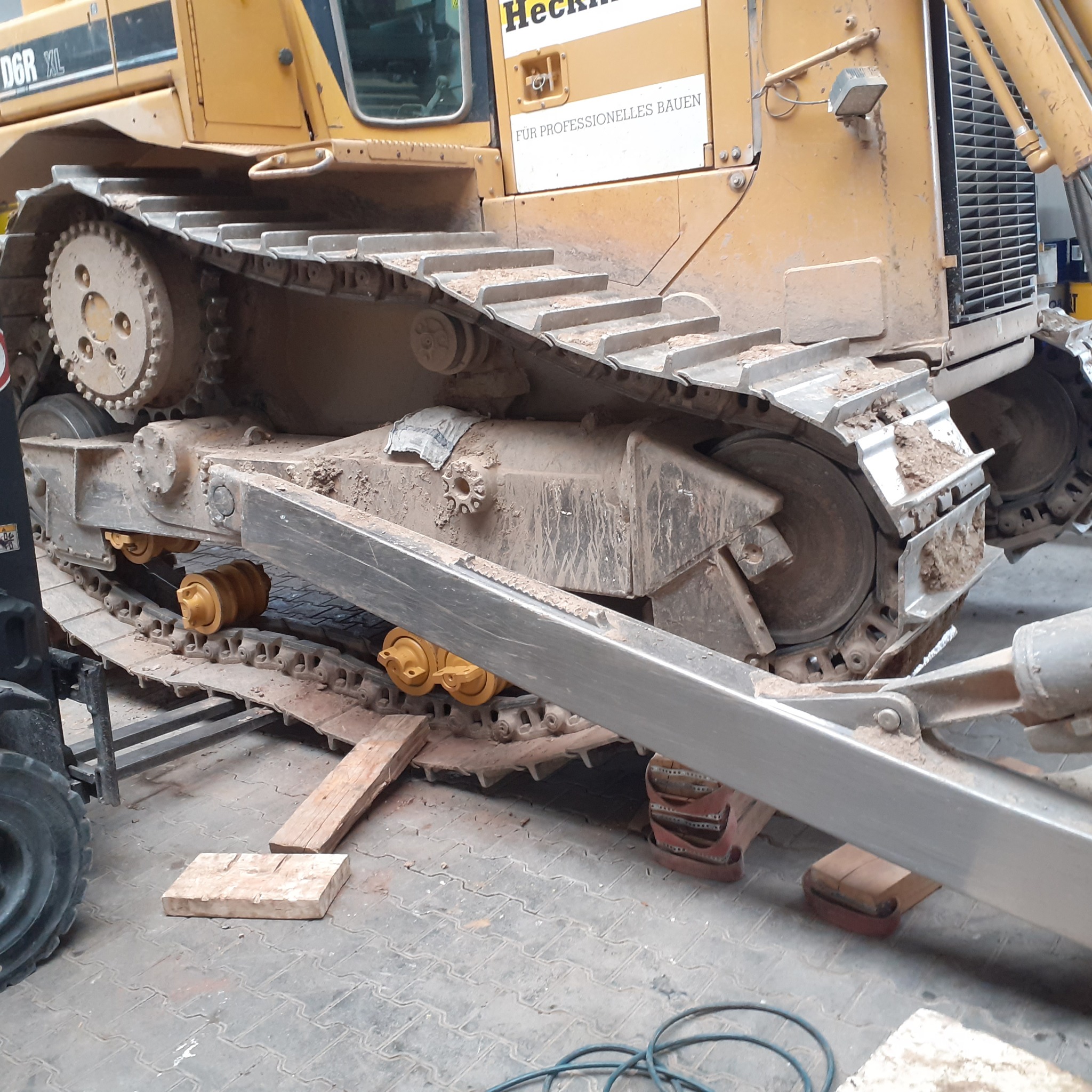 Gąsienica brudnej koparki D6R XL podczas serwisu w Lubinie, podparta drewnianymi klockami, z widocznymi elementami układu jezdnego i przewodami.