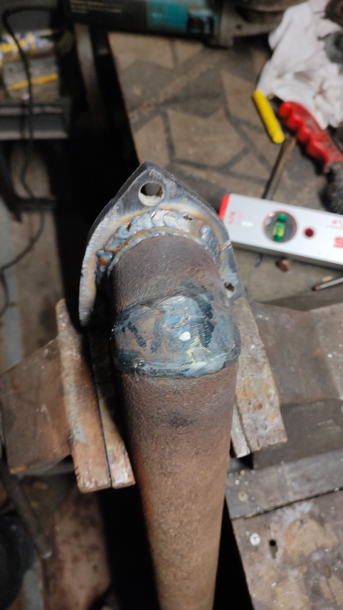 Rura wydechowa z przyspawanym kołnierzem, zamocowana w imadle, widoczne ślady spawania i rdzy, poziomica w tle.