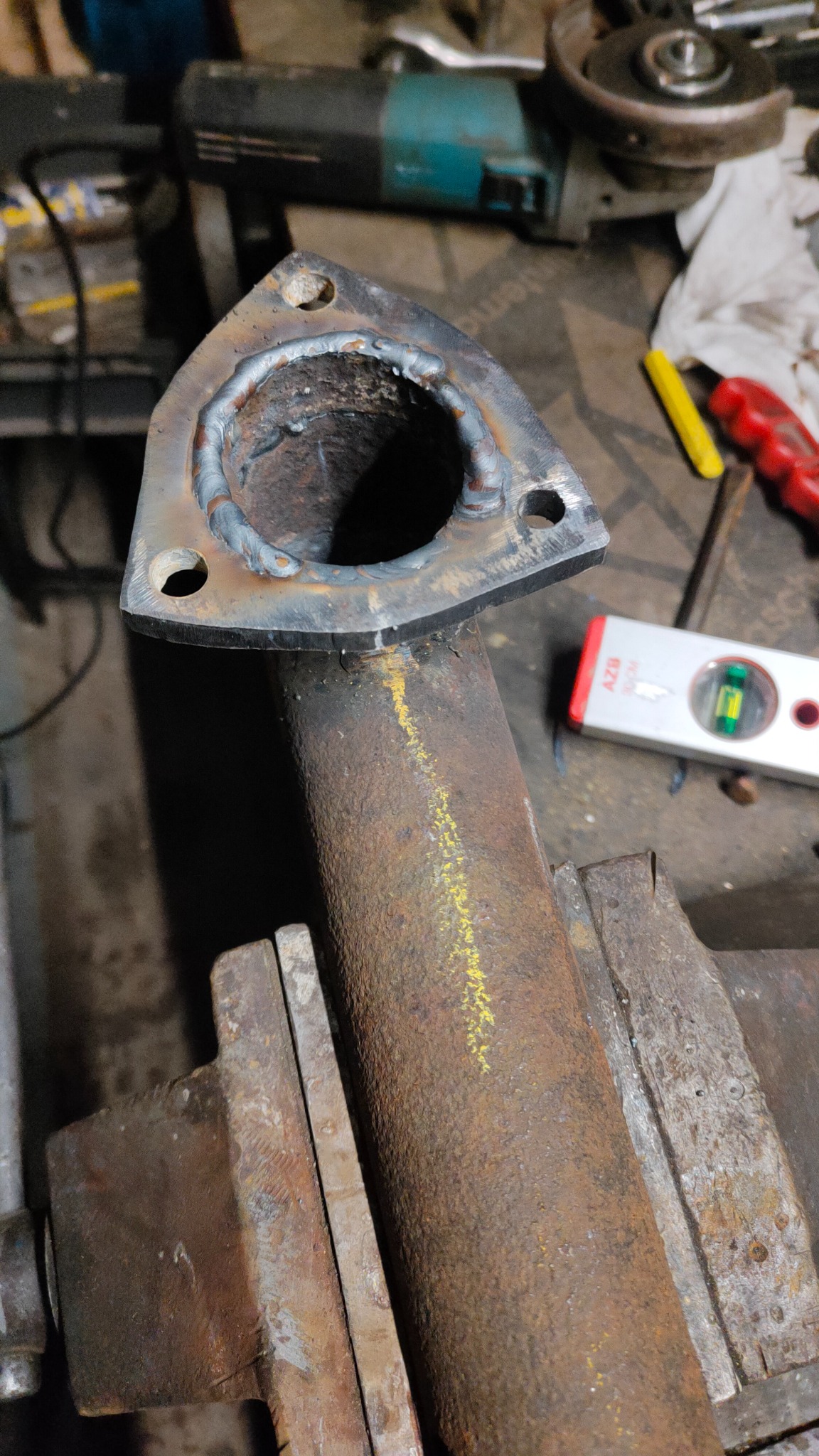 Fragment rury z przyspawanym kołnierzem, mocno zardzewiała, z żółtą linią zaznaczoną kredą, uchwycona w imadle warsztatowym, w tle narzędzia i poziomica.