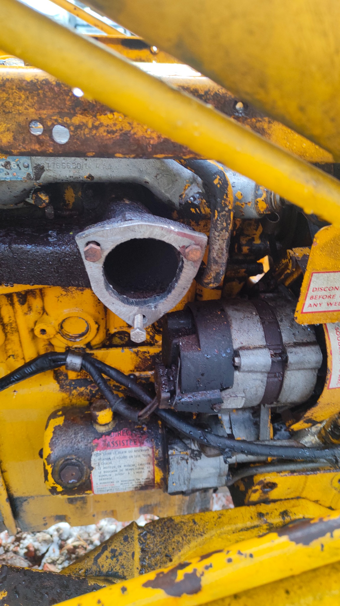 Żółta, zabrudzona olejem część silnika maszyny z widocznym przyłączem spalin, alternatorem i tabliczką informacyjną z tekstem w języku francuskim.