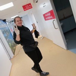 Karolina Szabelska - Uśmiechnięta kobieta w rękawiczkach ochronnych pozuje w korytarzu placówki medycznej, trzymając kubek po napoju i czarną czapkę.