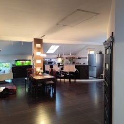 Karolina Szabelska - Przestronne wnętrze domu z otwartą kuchnią, jadalnią i salonem, widoczne meble, sprzęt AGD i akwarium, podłoga z ciemnego drewna, w tle jasne światło wpadające przez okno dachowe.