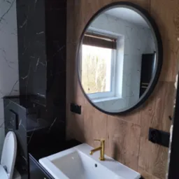 Nowoczesna łazienka z umywalką nablatową, złotą baterią, okrągłym lustrem i płytkami imitującymi drewno oraz marmur.