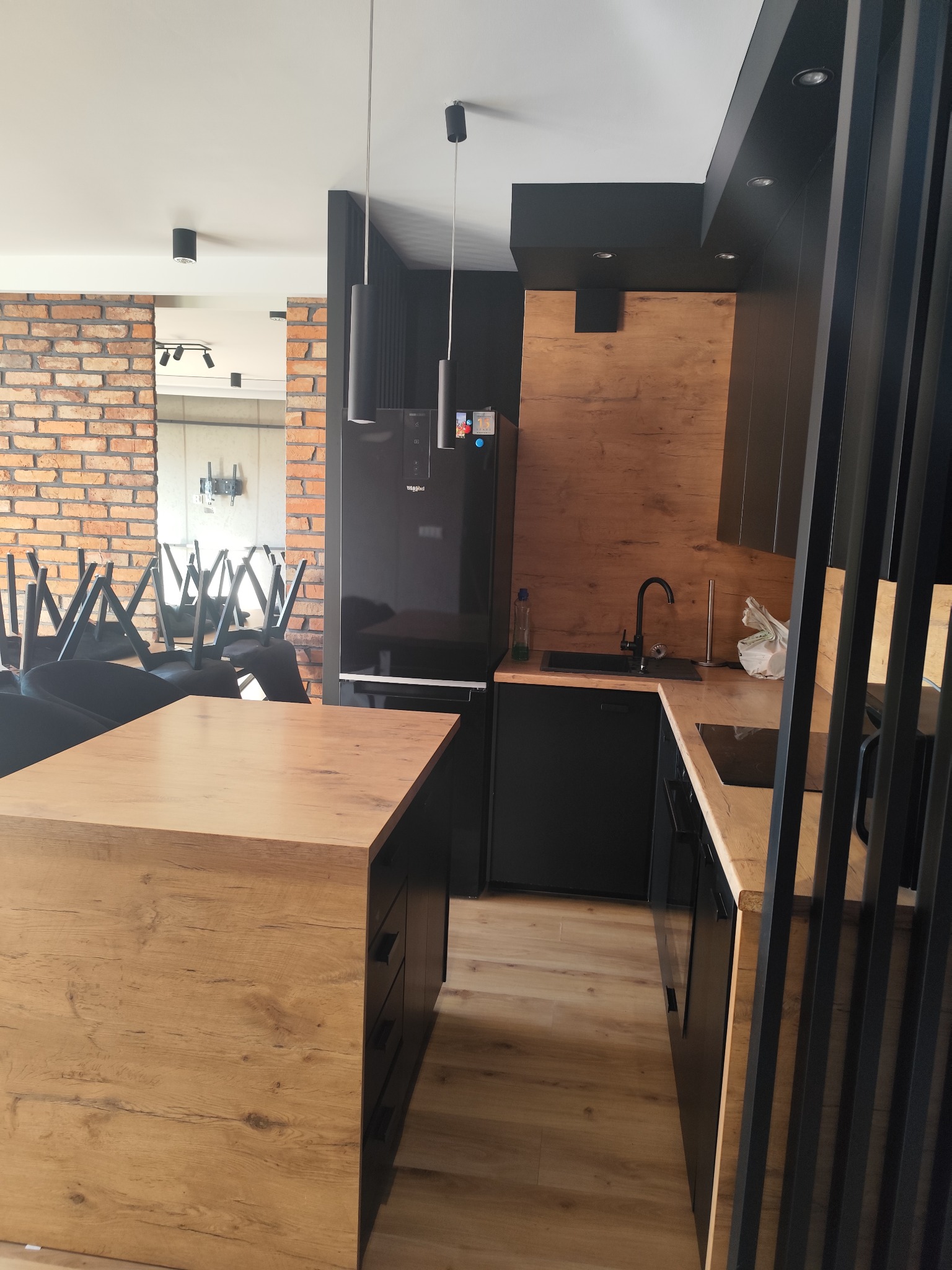 Nowoczesna kuchnia z czarnymi szafkami i drewnianymi blatami, widok na ceglaną ścianę w tle i odwrócone krzesła.