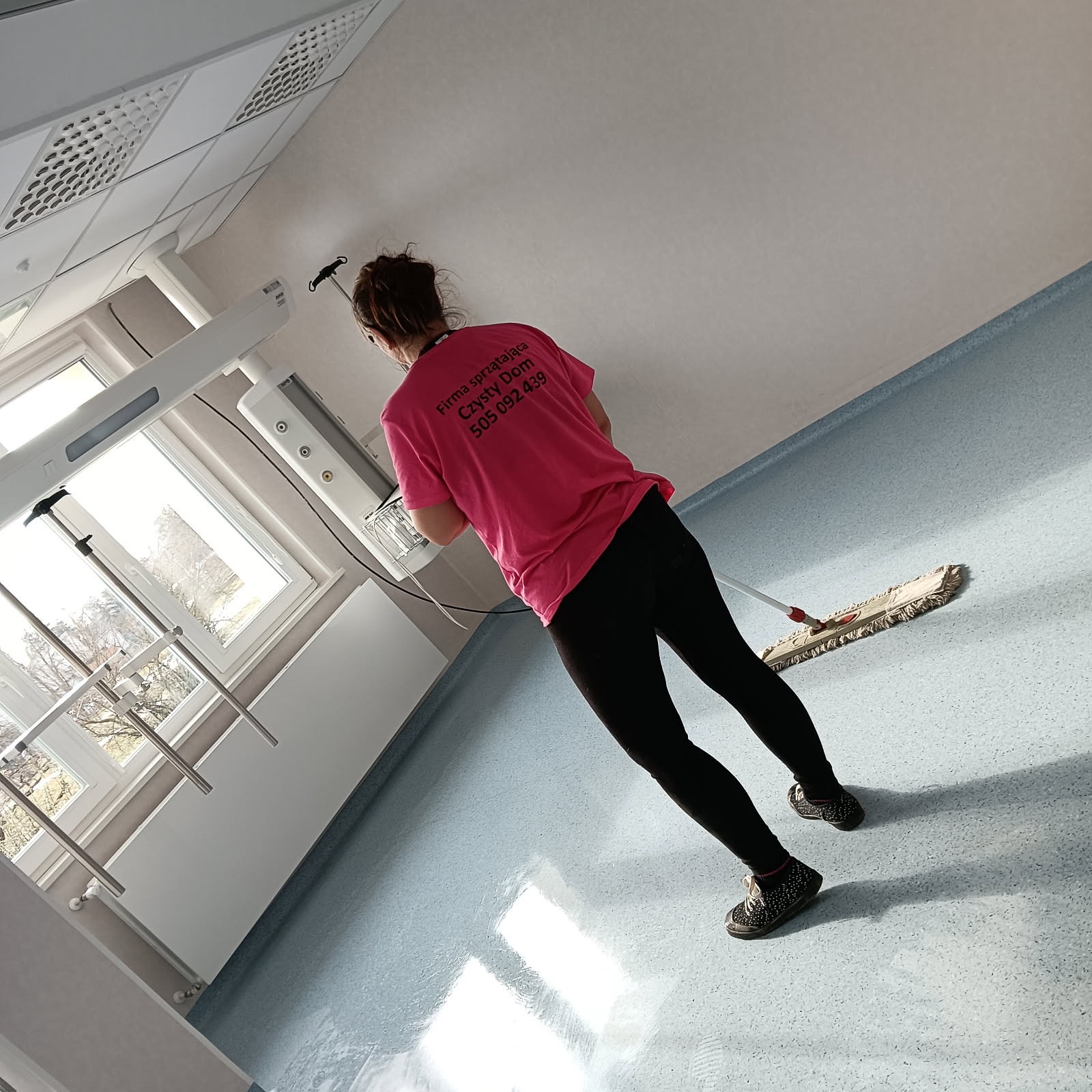 Pracownica w różowej koszulce z logo firmy sprzątającej myje podłogę mopem w pomieszczeniu szpitalnym z oknami i specjalistycznym sprzętem medycznym.