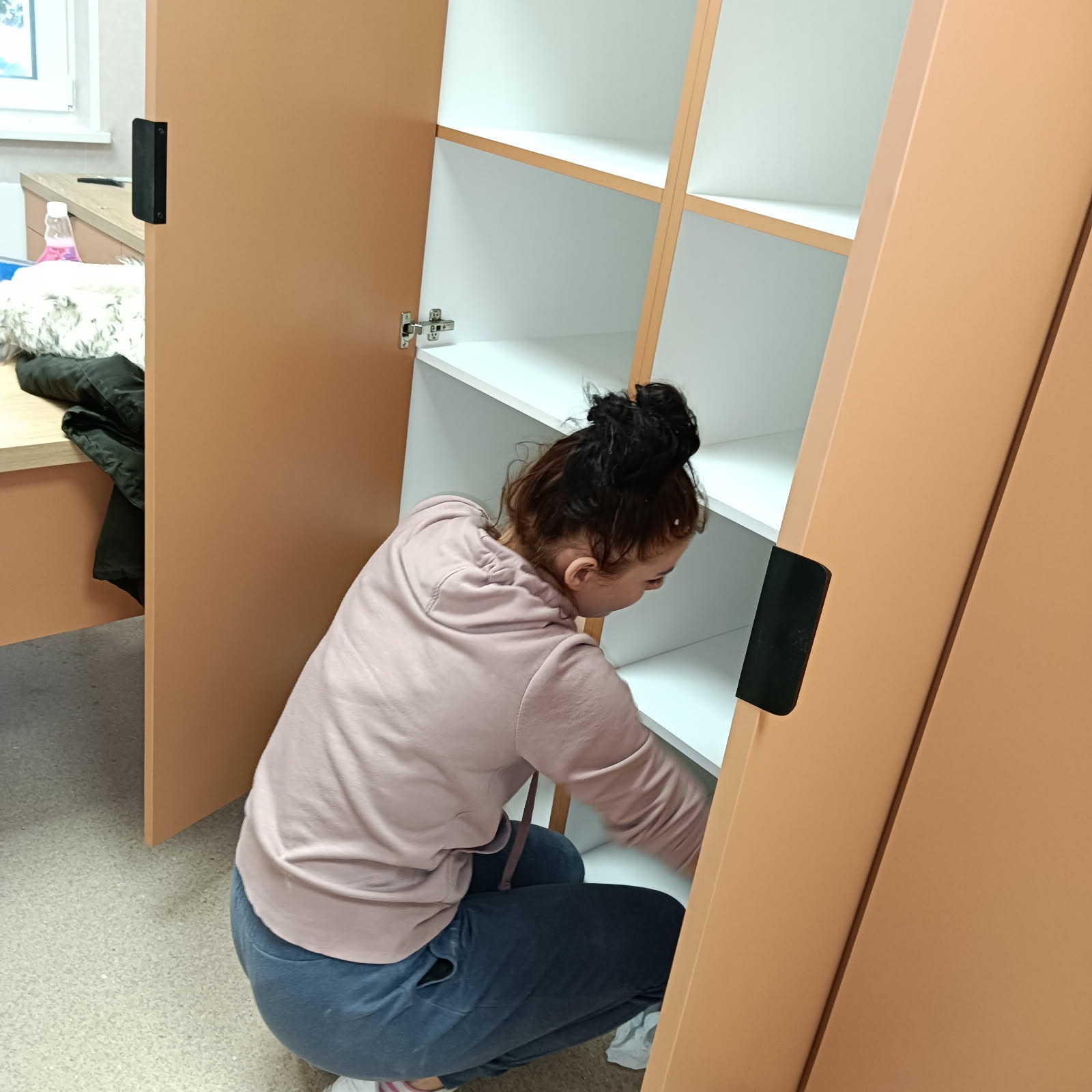 Kobieta w różowej bluzie i dżinsach czyści puste, białe półki w szafie o jasnobrązowych drzwiach, widoczne zawiasy i czarne uchwyty.