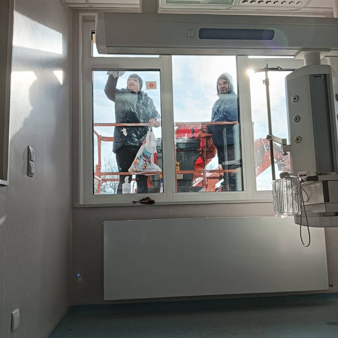 Dwaj pracownicy na podnośniku koszowym myją okna wewnątrz szpitalnego pomieszczenia z widoczną aparaturą medyczną.
