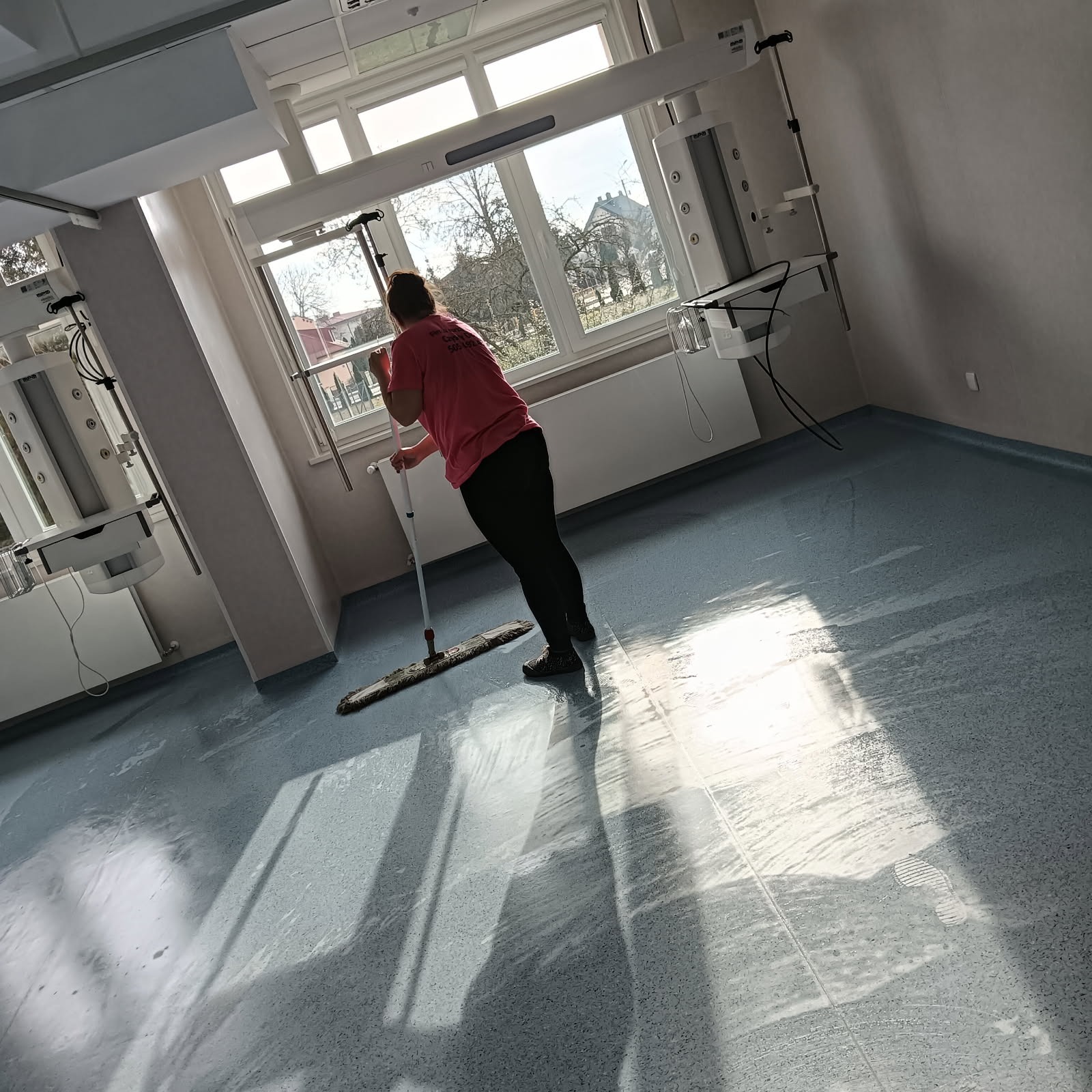 Pracownica w różowej koszulce mopuje podłogę w jasnym pomieszczeniu z dużym oknem w Warszawie. Na podłodze widoczne ślady wilgoci i odbicie światła słonecznego.