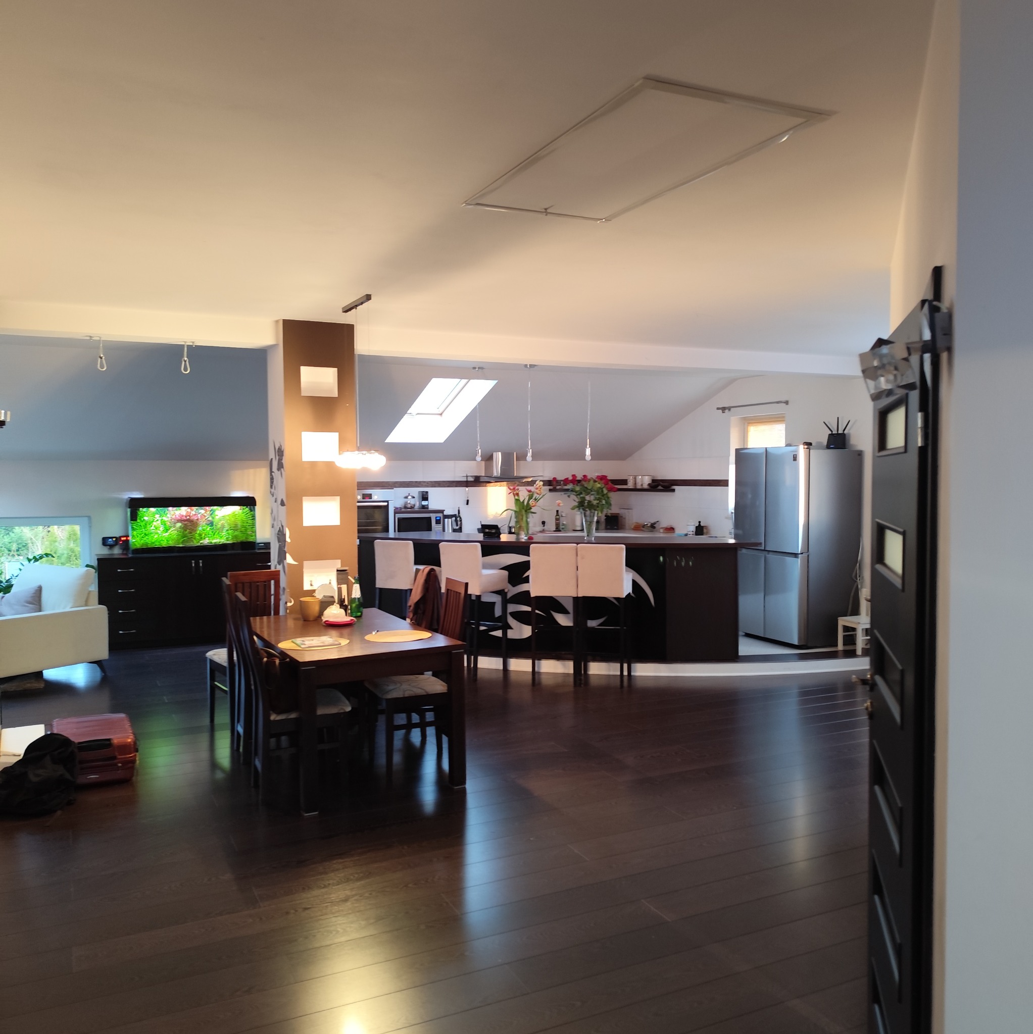 Przestronne wnętrze domu z otwartą kuchnią, jadalnią i salonem, widoczne meble, sprzęt AGD i akwarium, podłoga z ciemnego drewna, w tle jasne światło wpadające przez okno dachowe.