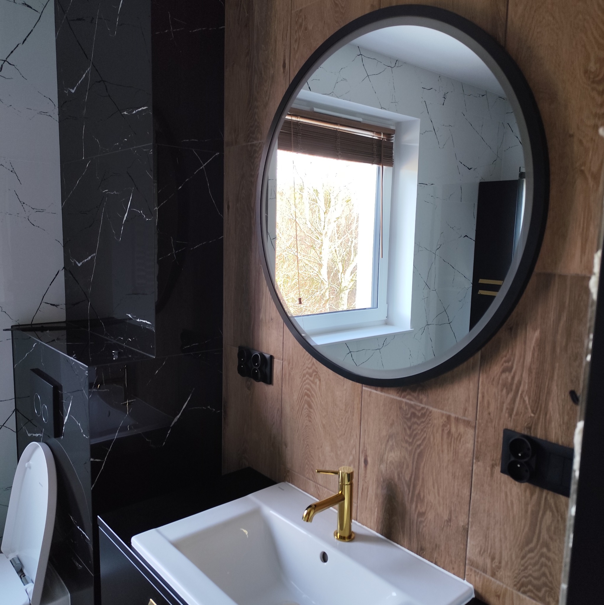 Nowoczesna łazienka z umywalką nablatową, złotą baterią, okrągłym lustrem i płytkami imitującymi drewno oraz marmur.