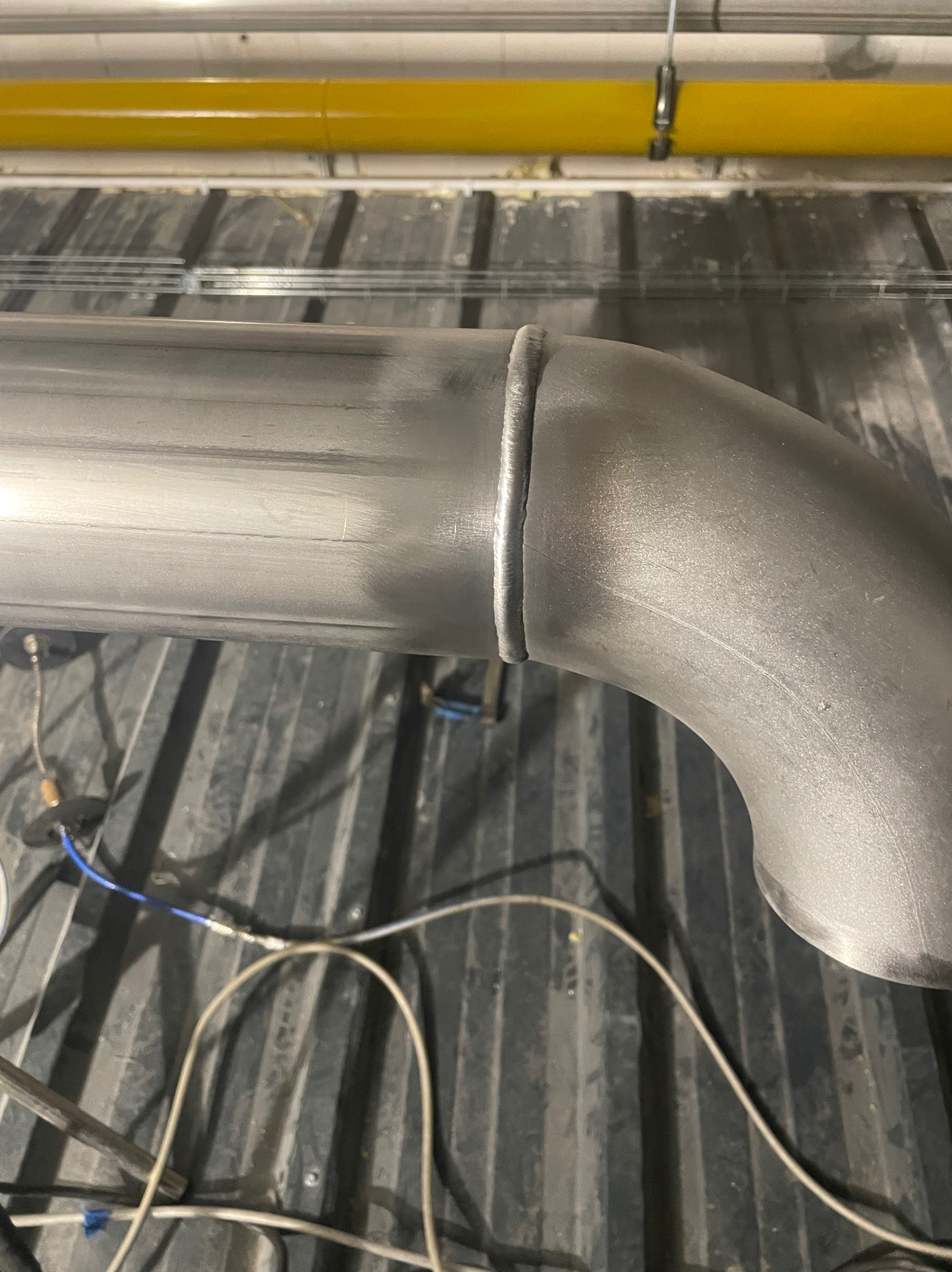 Szlifowany spaw na zakrzywionej rurze ze stali nierdzewnej, leżącej na ciemnej, żebrowanej powierzchni. Widoczne przewody elektryczne i żółta rura w tle.