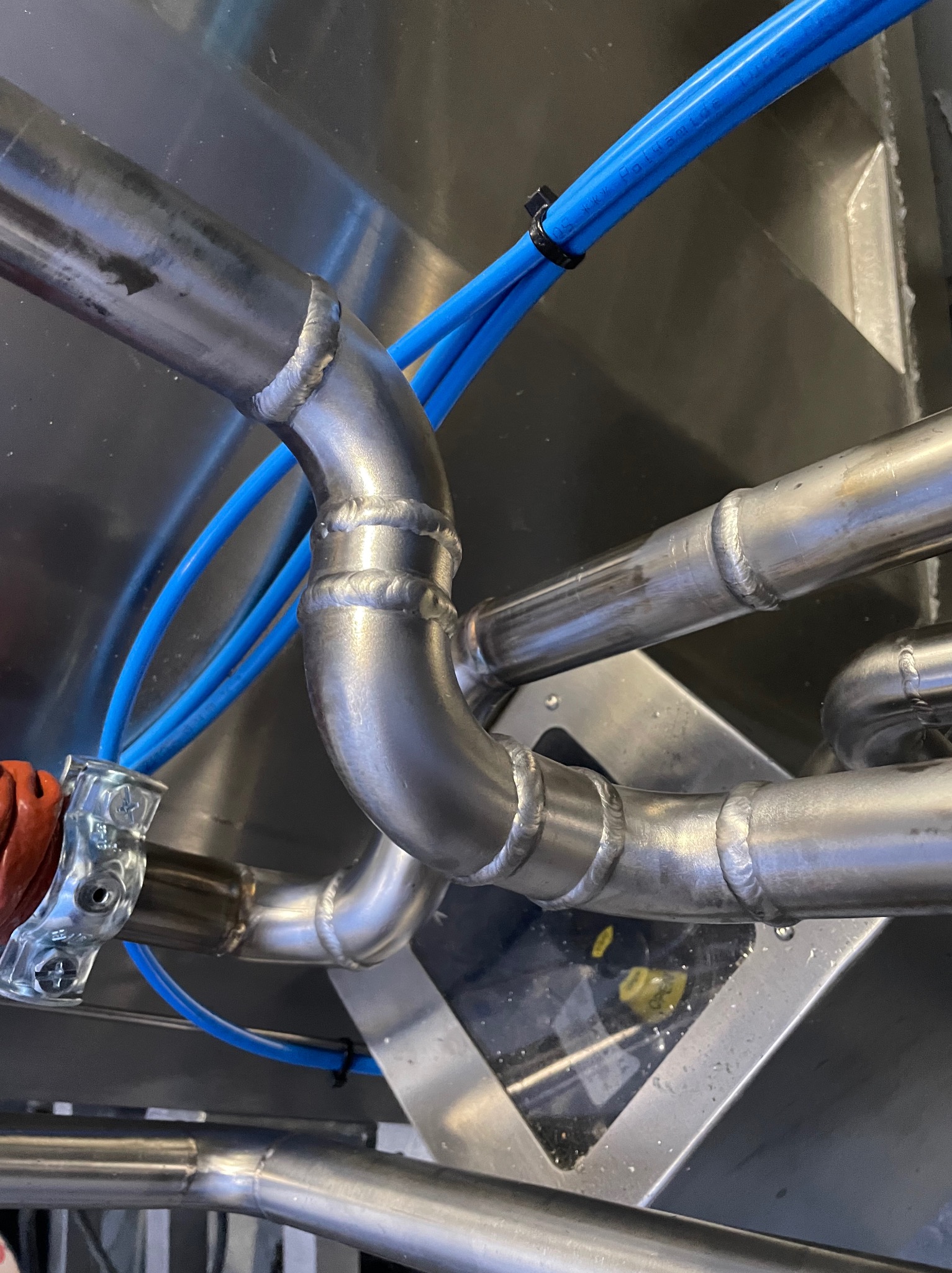 Zespawane rury ze stali nierdzewnej z niebieskimi przewodami, przymocowane do metalowej konstrukcji. Widoczne spoiny i elementy montażowe.