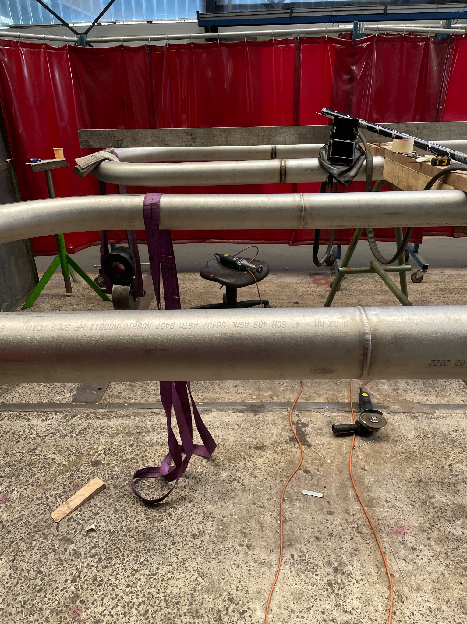 Spawane rury ze stali nierdzewnej na warsztacie, widoczne spoiny, narzędzia spawalnicze, pasy mocujące i oznaczenia na rurach. Czerwona zasłona w tle.