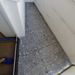 Andrii Rybchak - Szara, nakrapiana terakota ułożona na podłodze, z użyciem poziomujących klipsów i sznurka, widoczny fragment buta pracownika i fragment drzwi.