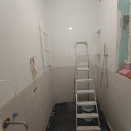 Andrii Rybchak - Wnętrze łazienki w trakcie prac wykończeniowych: białe płytki na ścianach, drabina malarska, częściowo zamontowana toaleta, widoczne instalacje hydrauliczne i fragment ściany z płyt...